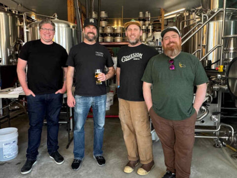 four people standing in a brewery in front of stainless steel brewing equipment