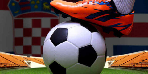 A soccer player’s orange boot on a football in a stadium with national flags in the background