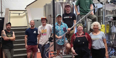 A group[ of people standing in a brewery, surrounded by brewing equipment