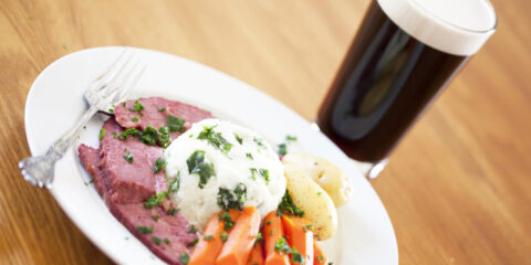 Two mugs of golden beer with foam and smoked meat pieces on a rustic wooden table beside a dark beer bottle.