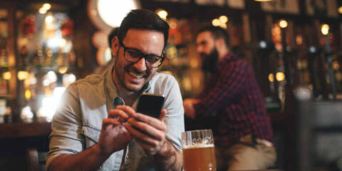 Download Tongits Games And Watch Your Wins Grow: Perfect For Craft Beer Fans Looking To Unwind Man relaxing in a bar, playing a game on his phone, drinking craft beer.