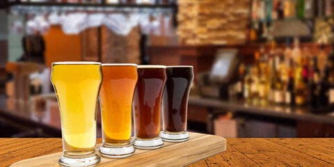 Four glasses of craft beer in a tasting flight, ranging from pale golden to dark brown, displayed on a wooden paddle at a modern bar.