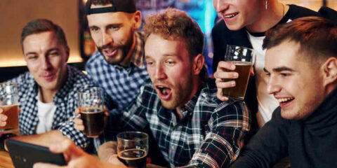 A group of five excited men huddles together at a bar, holding glasses of beer and reacting enthusiastically to something on a smartphone.