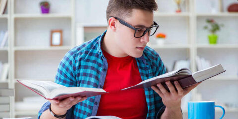 A young adult wearing glasses studies two open books at a desk.