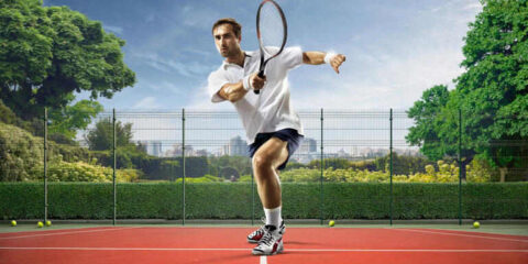 Male tennis player in action, hitting a forehand on an outdoor court, dynamic sports shot with racket motion, fenced court surrounded by greenery and city skyline in the background.