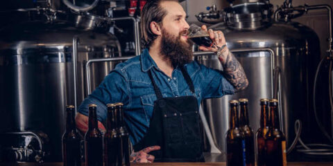 Bearded brewer sampling craft beer in a modern brewery, highlighting small-batch fermentation and hands-on brewing.