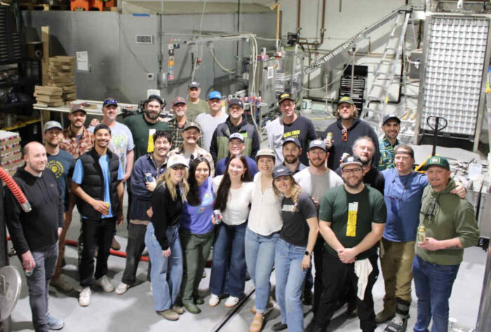 A group of brewers standing together in a brewery, surrounded by  stainless steel equipment