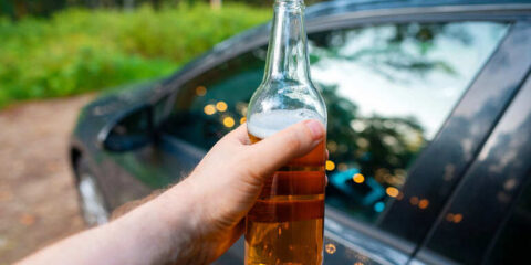 Hand showing a beer bottle close to a car, emphasizing the need to be safe and avoid driving while intoxicated on craft beer tours.