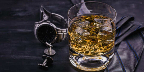 Glass of Scotch whisky with ice on a dark table beside a watch and tie.