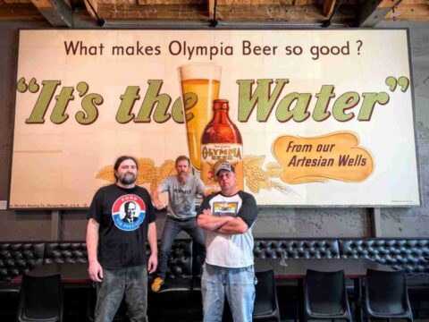 Three men standing in front of a historic Olympia Beer billboard