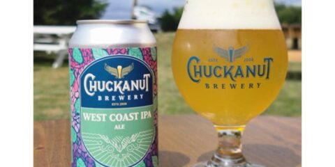 A can of beer next to a glass of beer, sitting on a table outside with grass and trees in the background