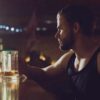 A man sits alone at a dimly lit bar, looking thoughtfully at a glass of alcohol.