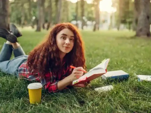 How College Students Can Balance Studying, Hobbies, And Social Life Without Burning Out College student girl relaxing in a garden with a book while balancing study, hobbies, and social life.