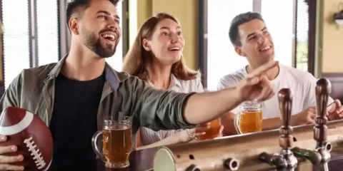 Friends enjoying beer and watching football at a sports bar while discussing parlay bets on game day.