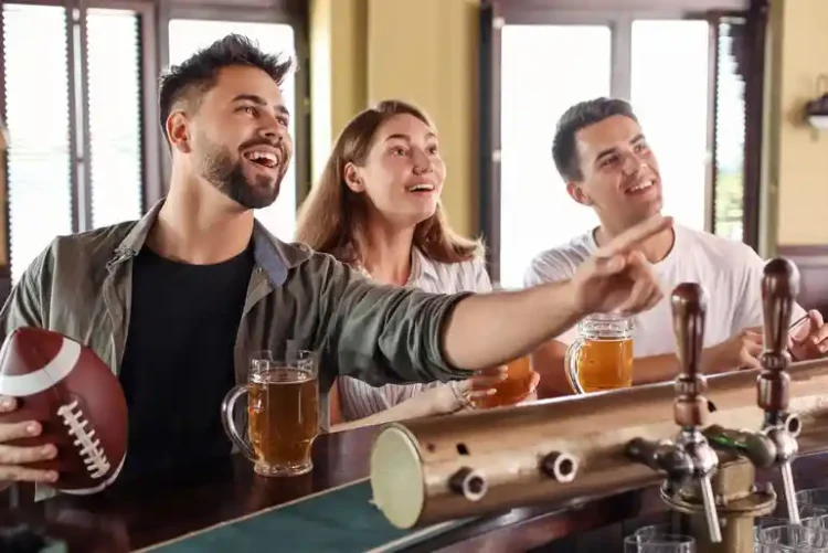 Friends enjoying beer and watching football at a sports bar while discussing parlay bets on game day.
