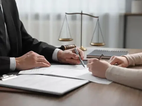 A lawyer in a black suit pointing at legal documents while consulting with a client at a desk, featuring a scale of justice in the background.