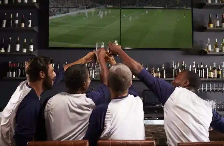 The Pint Is No Longer the Whole Night Friends raising beer glasses while watching a soccer match together at a lively bar, highlighting the social experience beyond the pint.
