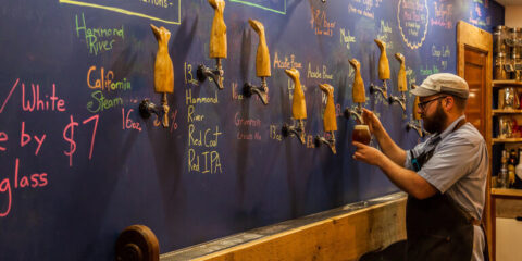 A taproom with a cozy interior, featuring a bartender standing in front of a blue board.