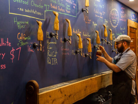 The Washington Bottle-Share Playbook: Small Pours, Local Stories and a Table Game Between Flights A taproom with a cozy interior, featuring a bartender standing in front of a blue board.