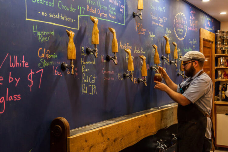 A taproom with a cozy interior, featuring a bartender standing in front of a blue board.