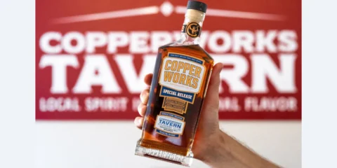 Copperworks Tavern Nears Opening, Join the Founders Club Now A person holding a bottle of Copperworks whiskey in front of a sign for the Copperworks Tavern