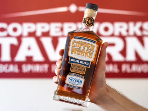 Copperworks Tavern Nears Opening, Join the Founders Club Now A person holding a bottle of Copperworks whiskey in front of a sign for the Copperworks Tavern