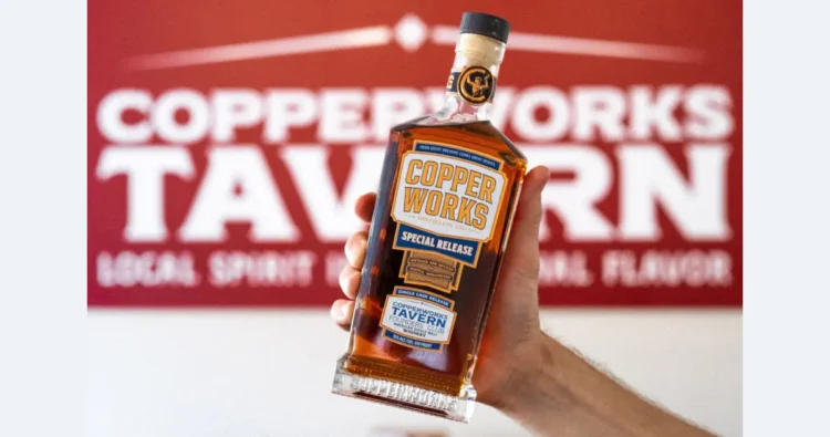 A person holding a bottle of Copperworks whiskey in front of a sign for the Copperworks Tavern