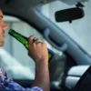A man sitting in the driver's seat of a car drinking a beer from a green bottle, illustrating drunk driving.