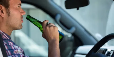 The Legal and Financial Consequences of a Drunk Driving Accident A man sitting in the driver's seat of a car drinking a beer from a green bottle, illustrating drunk driving.