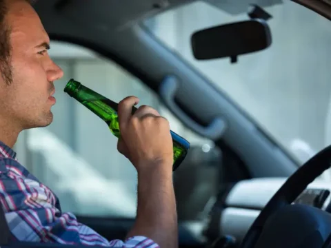 The Legal and Financial Consequences of a Drunk Driving Accident A man sitting in the driver's seat of a car drinking a beer from a green bottle, illustrating drunk driving.