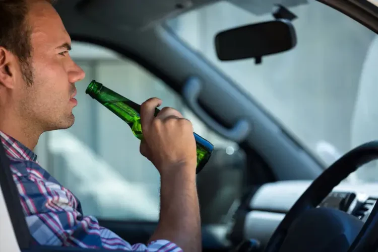 The Legal and Financial Consequences of a Drunk Driving Accident A man sitting in the driver's seat of a car drinking a beer from a green bottle, illustrating drunk driving.