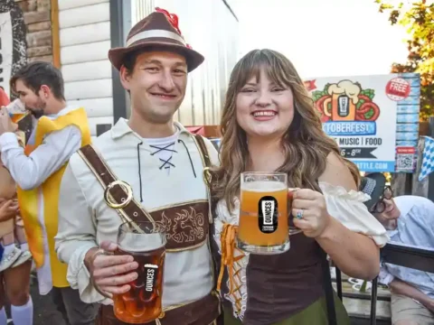 The Weekend’s Beer Events – October 2nd Through 4th, 2026 two people in Oktoberfest garb hoisting mugs of beer